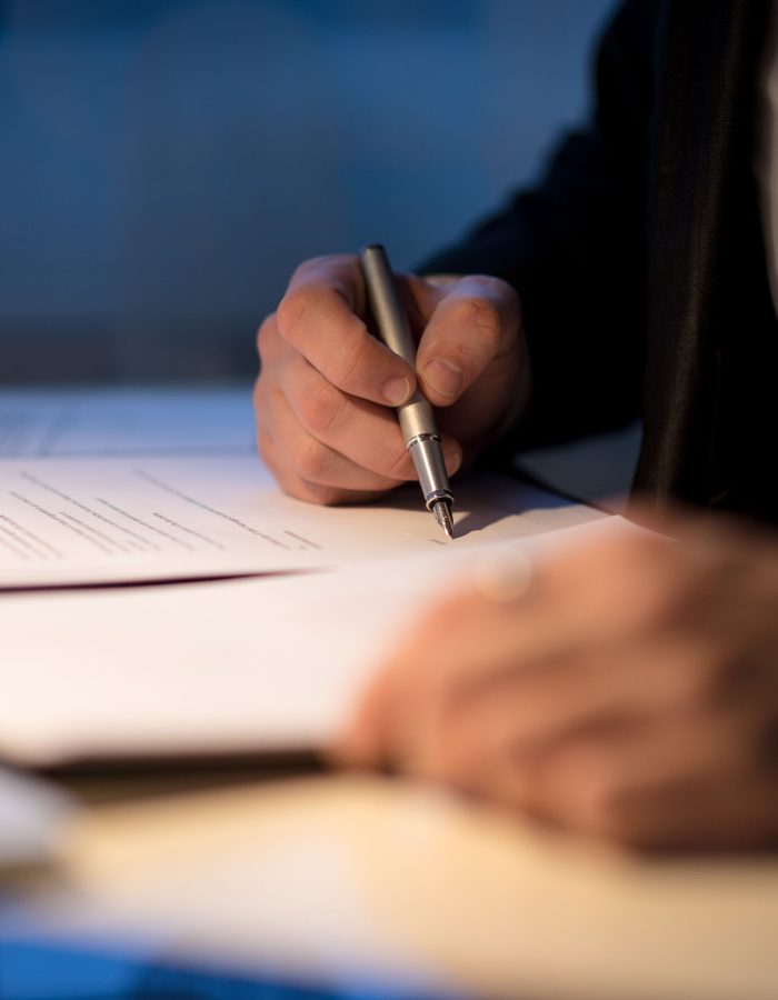 Businessman working late signing a document or contract in a dark office with a fountain pen by the light of a lamp, close up view of his hands.